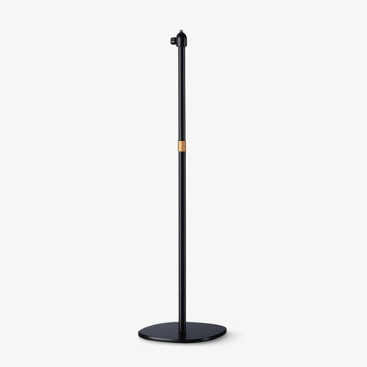 Product photograph of a minimalist matte black floor stand featuring a slim cylindrical pole, a prominent copper accent band, and a sleek oval base with an adjustable top mount